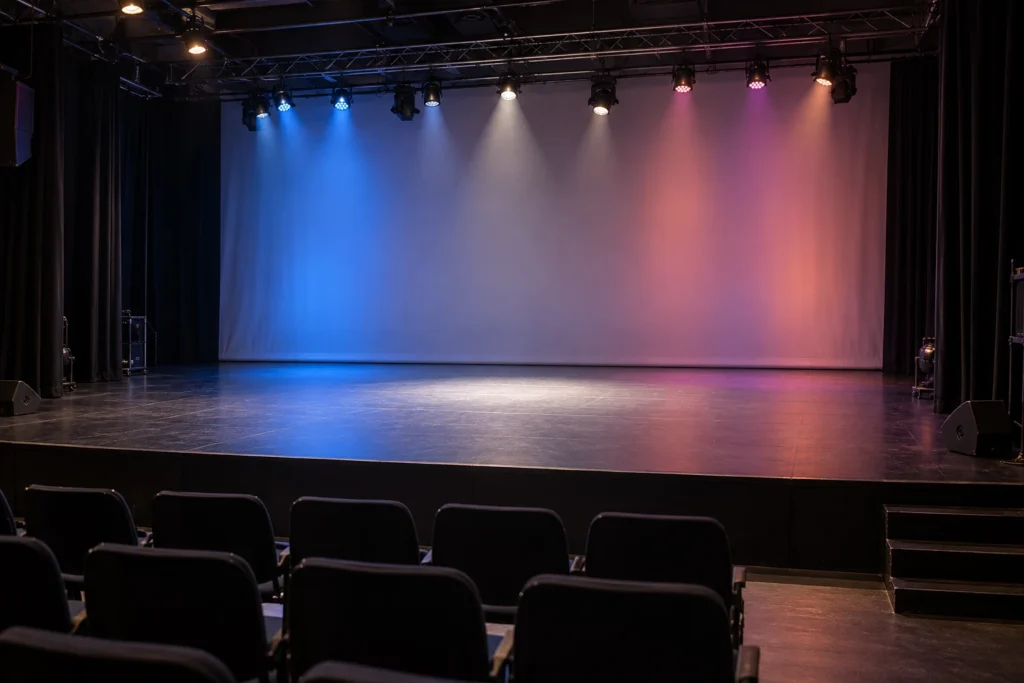 Empty theatre stage with blue and pink light on a grey backdrop, a white pool of light at centre stage and dark audience seating.